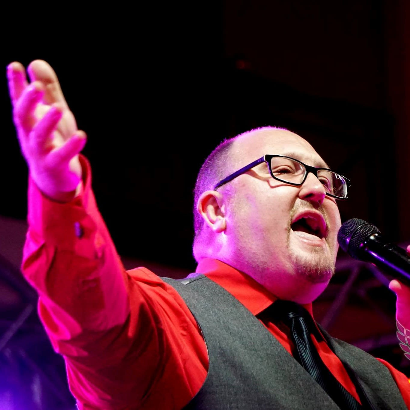 Jon paul cabaret party entertainer wearing red shirt and singing into microphone with right arm outstretched.