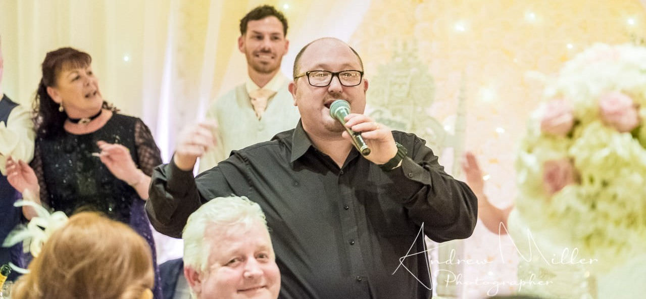 Groom and mother smile in background as jon paul singing waiter in black shirt sings by wedding guests in front.