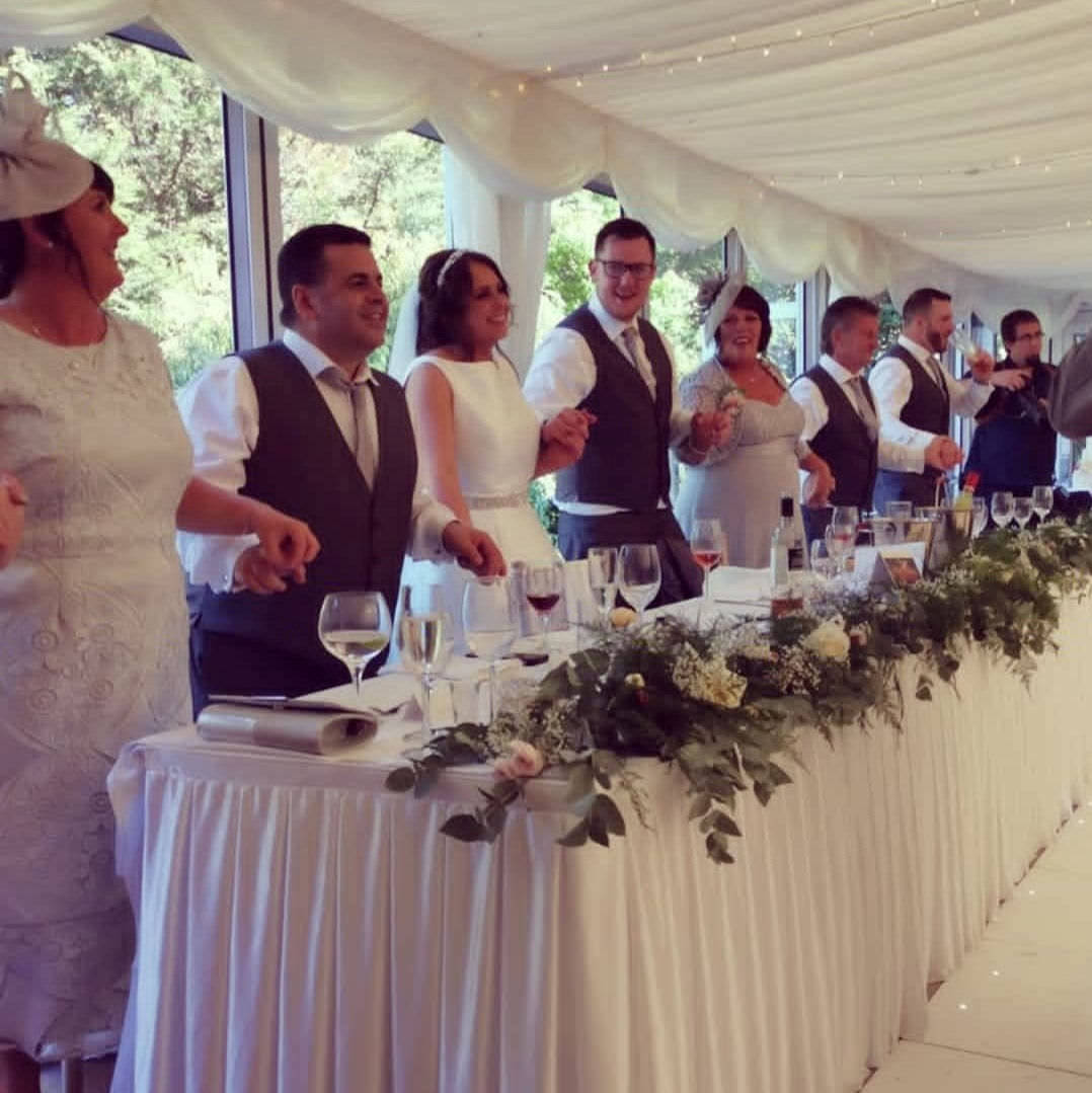 Family stand holding hands and sing along joyfully behind top table for jon paul singing waiter at wedding in south wales.