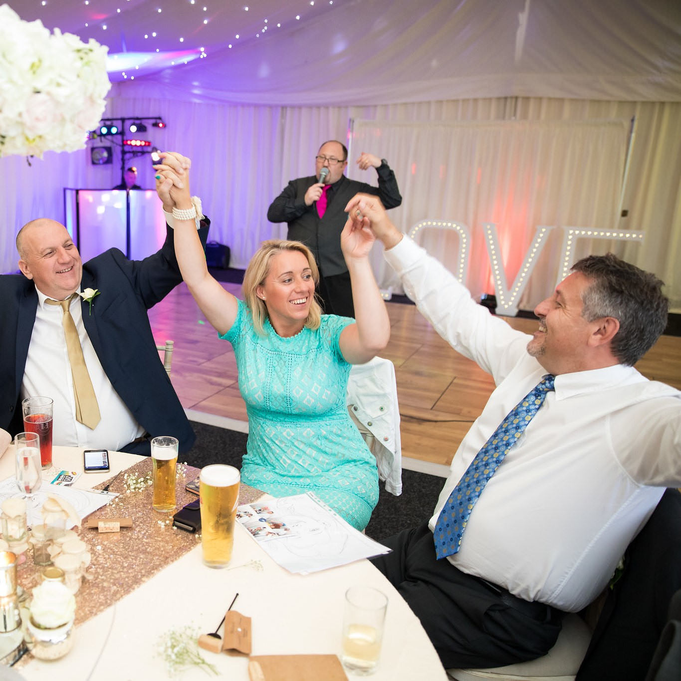 Guest at table clasp each other hands in the air as jon paul surprise singer sings behind them.