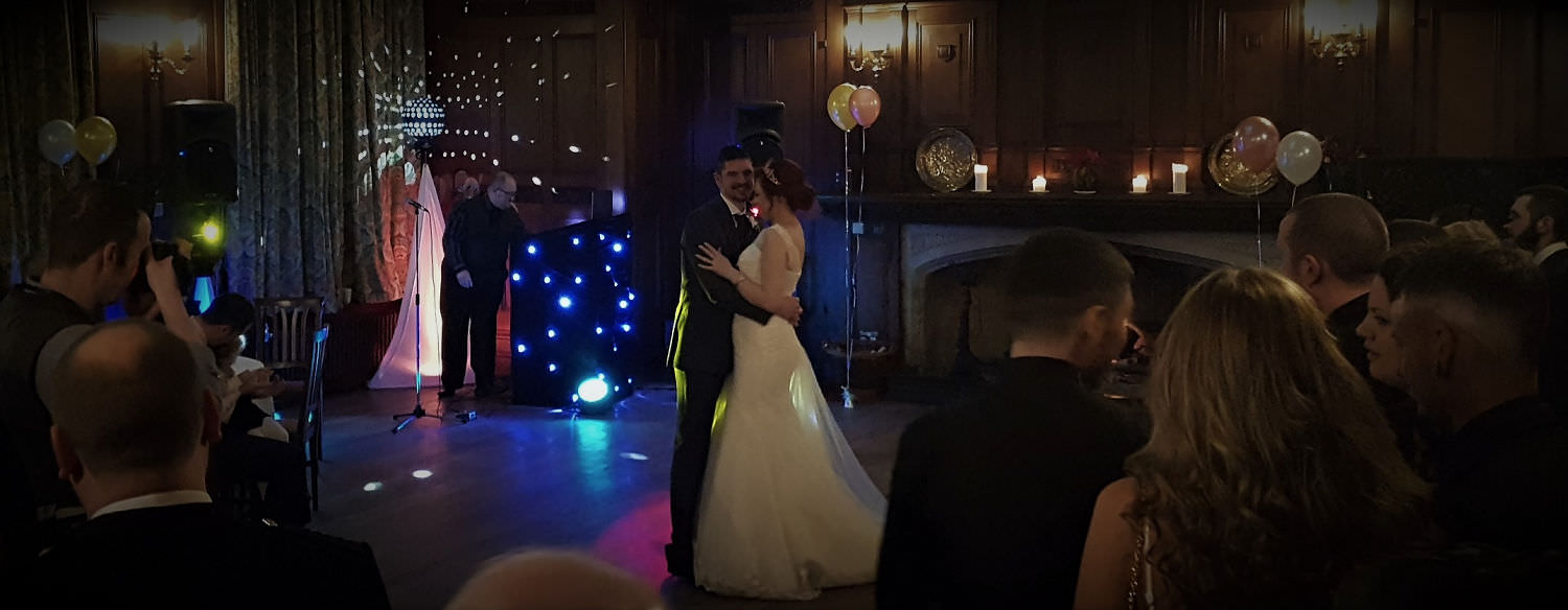 Newlyweds embrace in live first dance, with jon paul wedding singer next to twinkling DJ booth and lighting in the background.