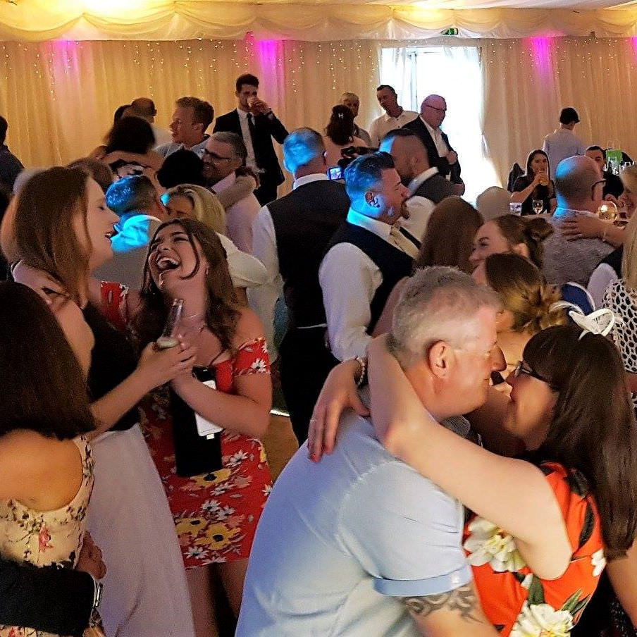 Full dance floor of dancing guests in pretty summer clothes laugh and embrace during slow song at wedding near bristol.