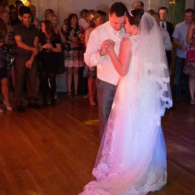 Bride and groom embrace tenderly in their first dance while family and friends gather around them to watch.
