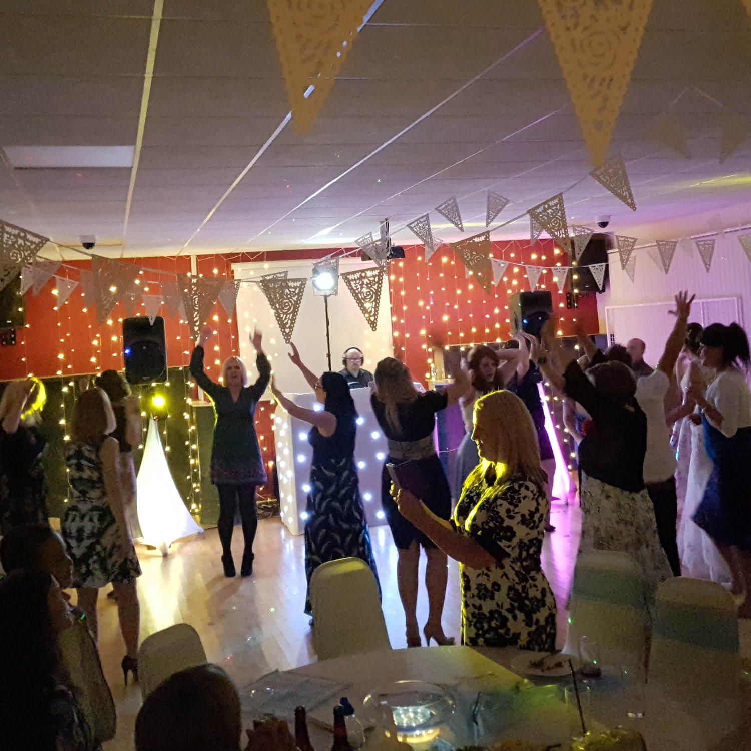 Guests raise their arms in air dancing in front of white DJ booth with lace bunting above their heads.