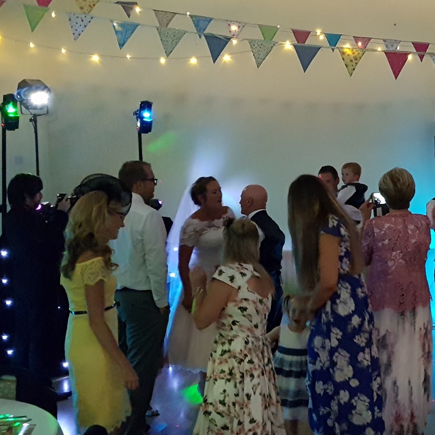 Wedding guests in summer clothes and multi-coloured bunting overhead surround bride dancing in front of disco lightshow.