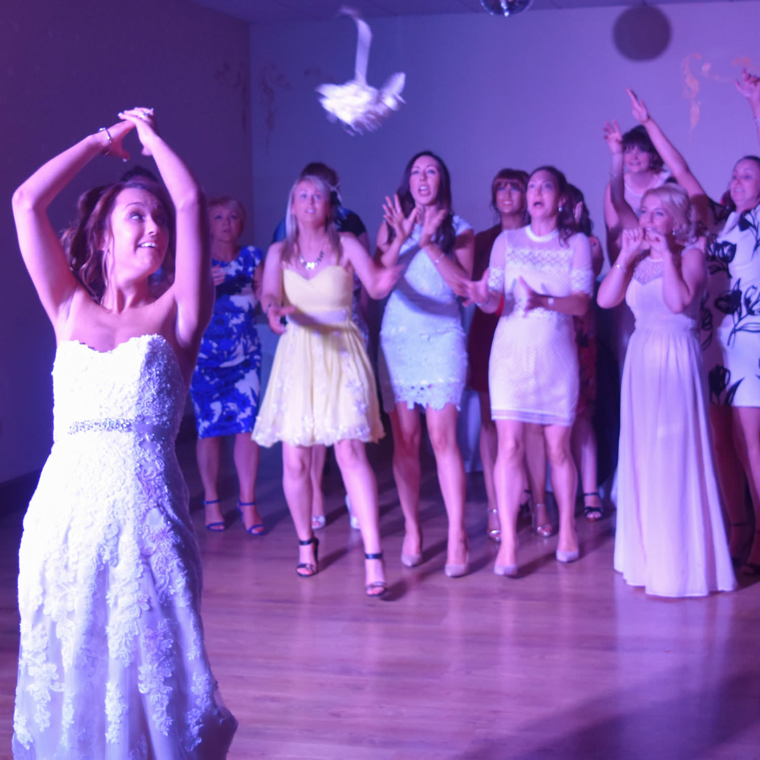 Bride throws bouquet over her shoulder to hen group bathed in disco lighting at wedding party near Bristol.